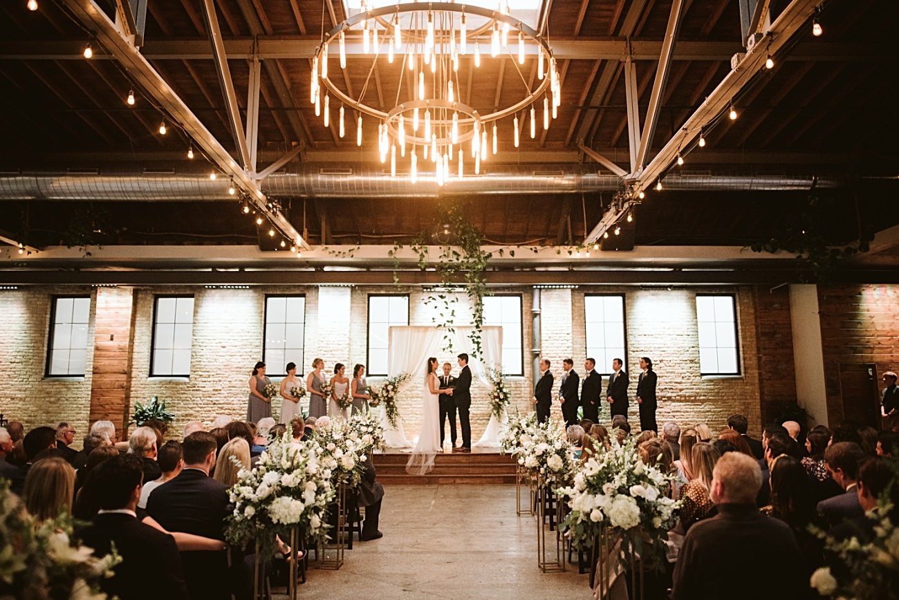 A bride and groom stand at the altar in an industrial-style venue, surrounded by their wedding party. Guests are seated in rows with white floral arrangements along the aisle under warm, hanging lights—a true gem among Wisconsin Wedding Venues.