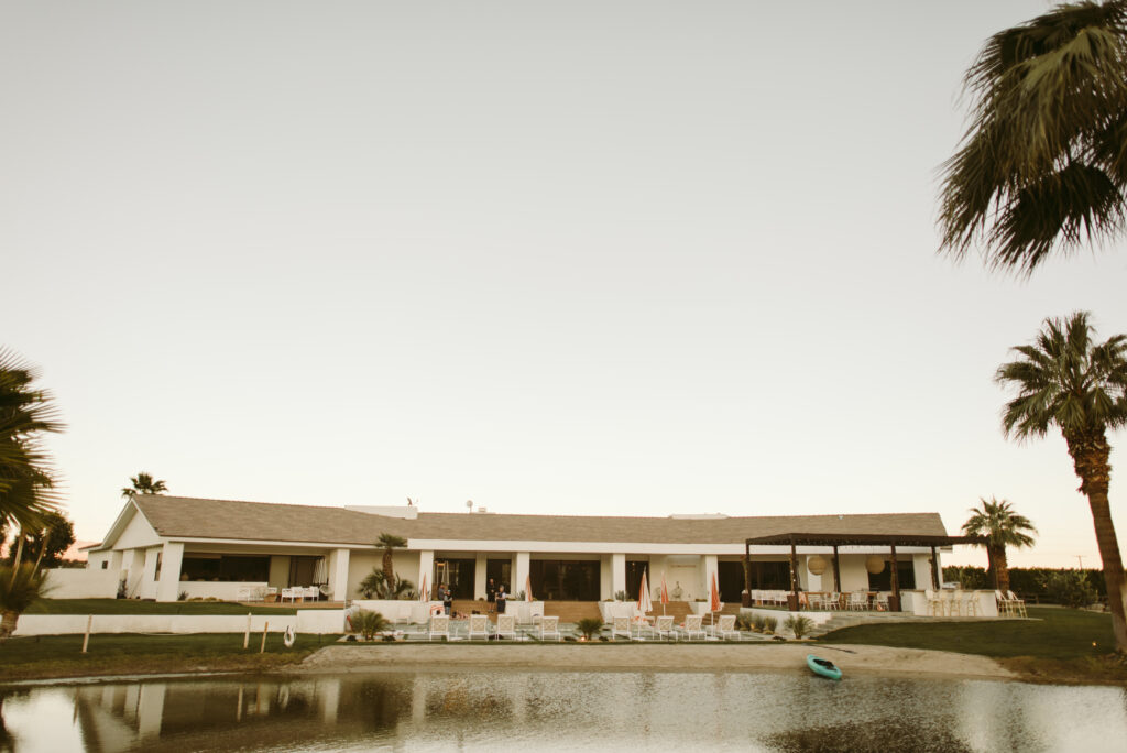 A modern, single-story house with a large patio faces a small pond—an ideal elopement destination—surrounded by palm trees and outdoor seating under a clear sunset sky. A blue kayak sits near the water’s edge.