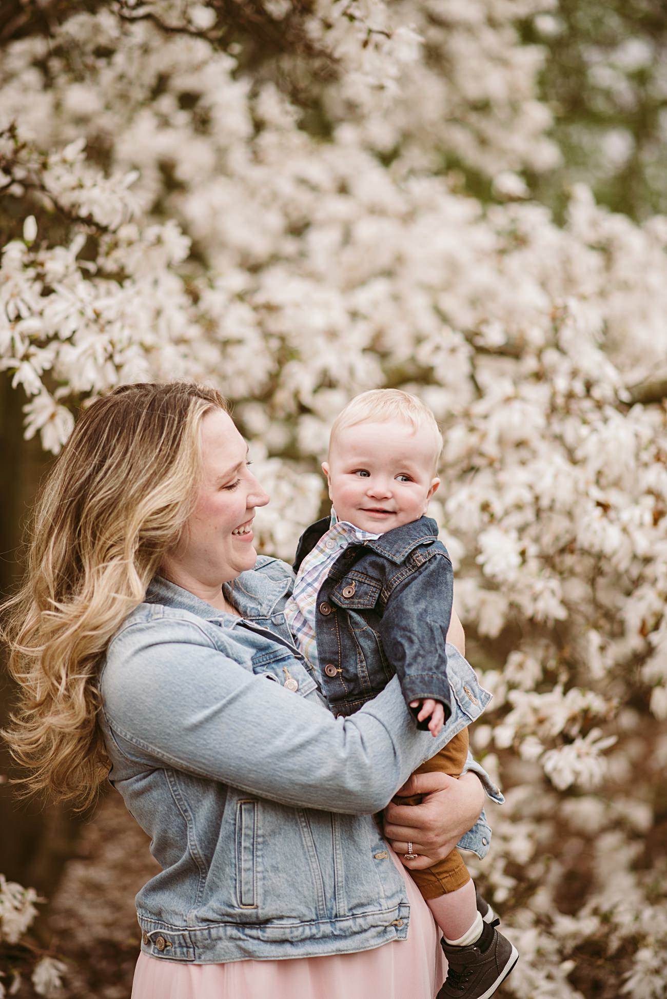 A woman with long blonde hair smiles while holding a baby in her arms. Both wear denim jackets and stand before blooming white flowers—a perfect scene of Spring Family Photography. The baby looks off to the side, appearing content.