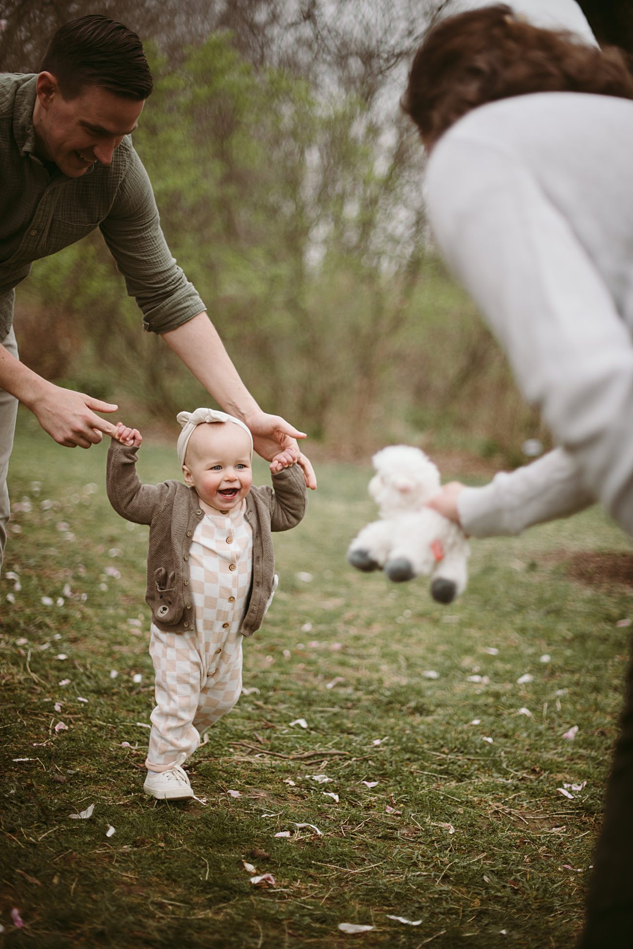 A smiling toddler walks on grass, holding an adult’s hands, while another adult holds a stuffed animal out to the child. The scene is outdoors with trees and greenery in the background.