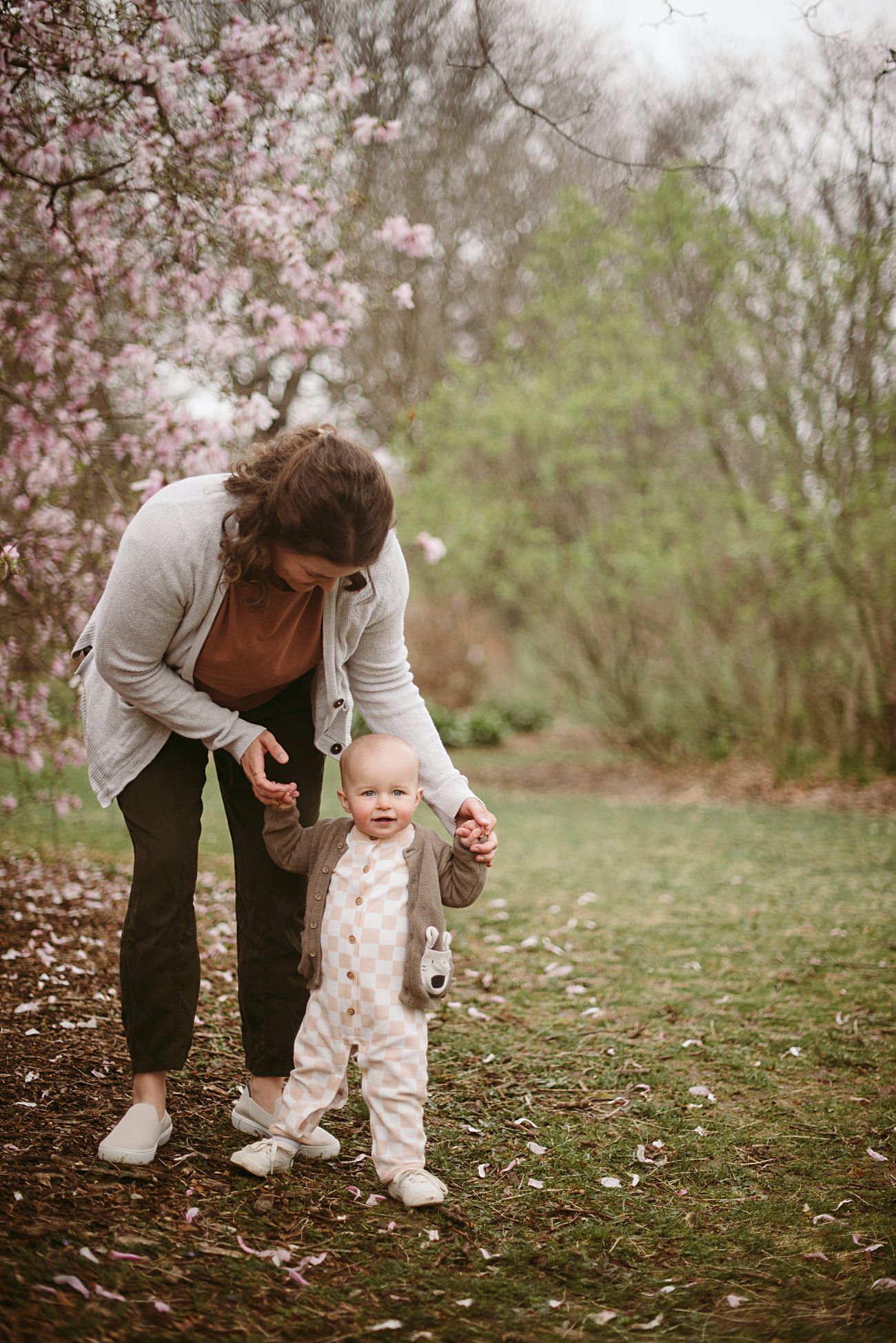A woman bends down to help a smiling baby stand and walk on a grassy path in a park, surrounded by blooming trees and scattered flower petals.