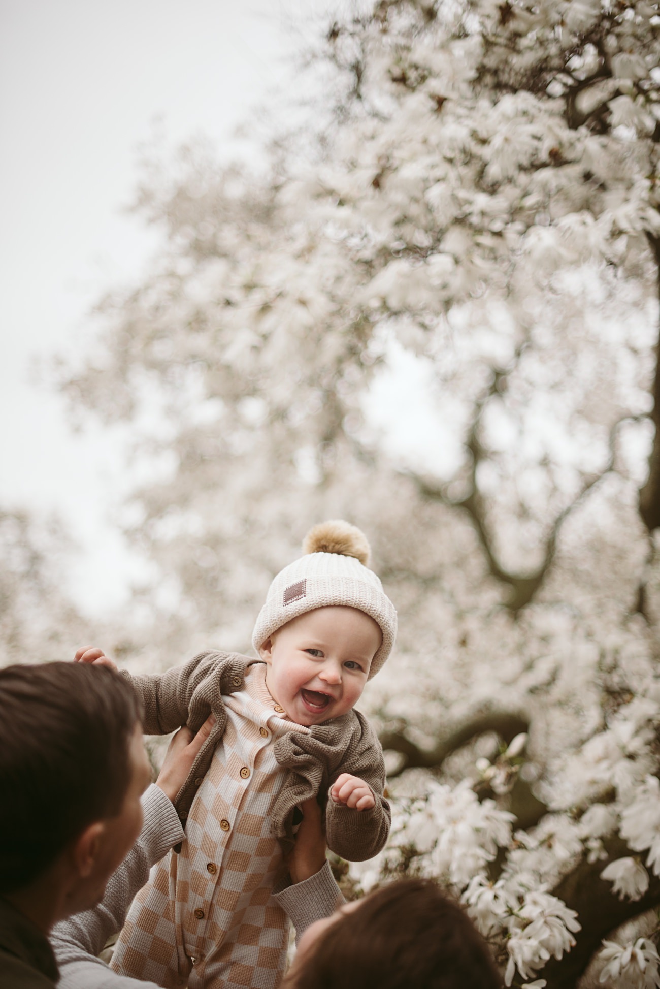 A happy baby in a knit hat and checkered outfit is lifted up by adults under blossoming trees with white flowers, smiling joyfully at the camera.