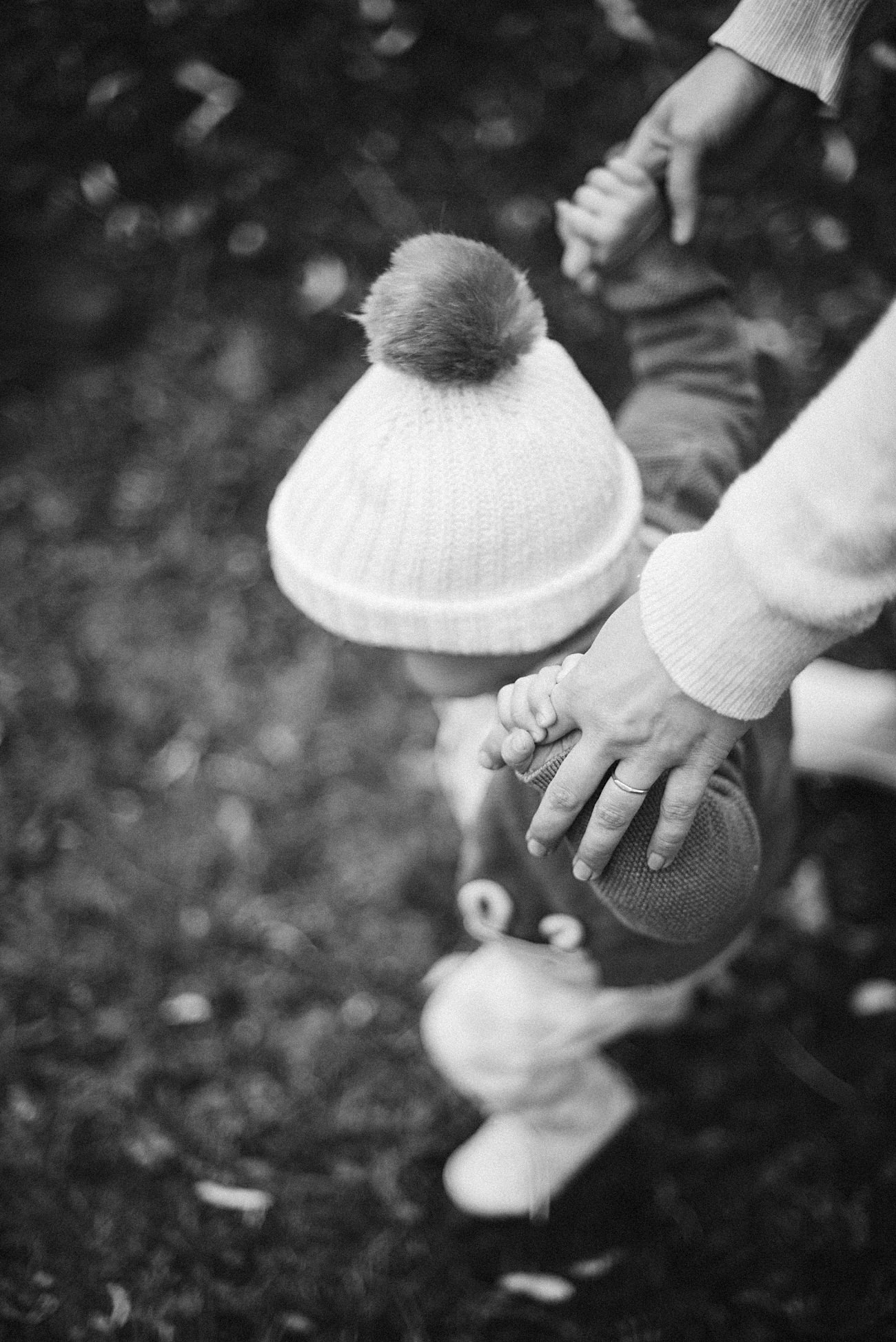 A black and white photo of a small child wearing a knit hat with a pom-pom, holding an adult’s hand for support while walking on grass. The adult’s hand gently steadies the child.