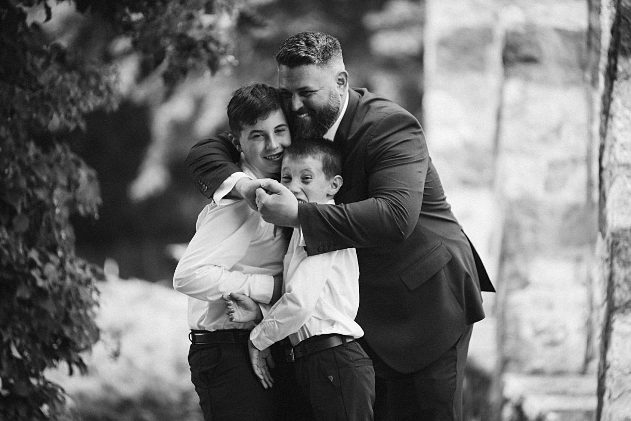 A man in a suit hugs two smiling boys, also dressed formally, in an outdoor setting with stone walls and greenery—captured in black and white as part of a 2025 wedding recap.