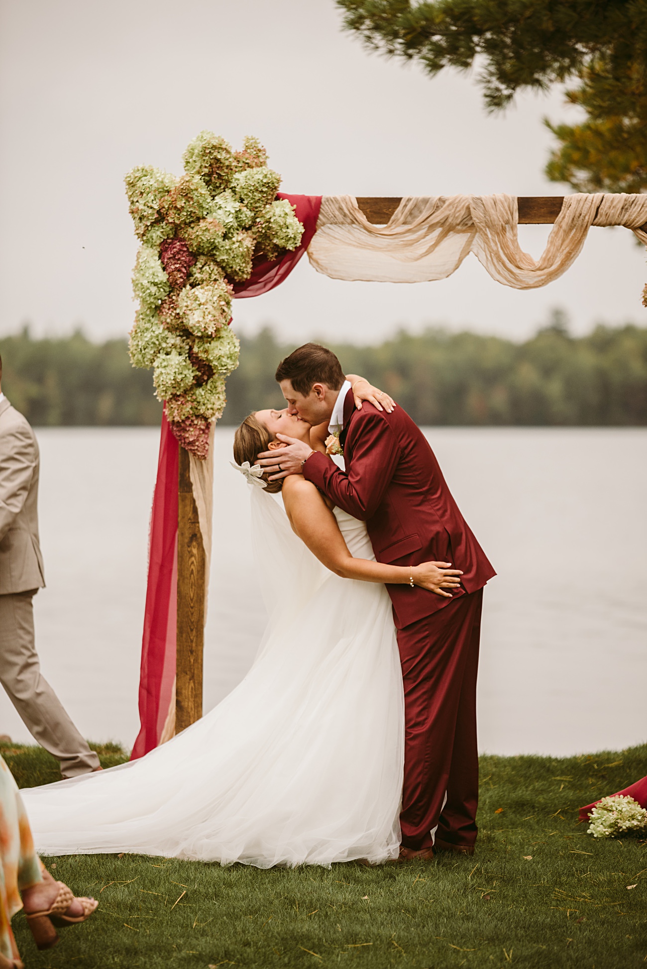 A bride and groom share a kiss under a decorated wooden arch by a lake, surrounded by greenery. The bride wears a white dress and veil, and the groom wears a burgundy suit in this unforgettable 2025 wedding recap.