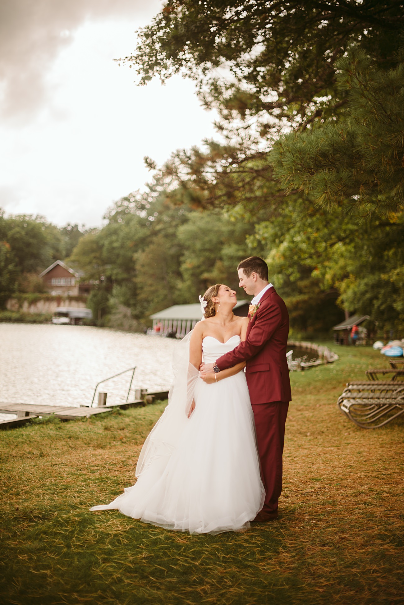 A bride in a white gown and a groom in a burgundy suit stand smiling at each other by a lakeside, surrounded by trees and rustic cabins—a picture-perfect scene for a romantic 2025 Wedding.