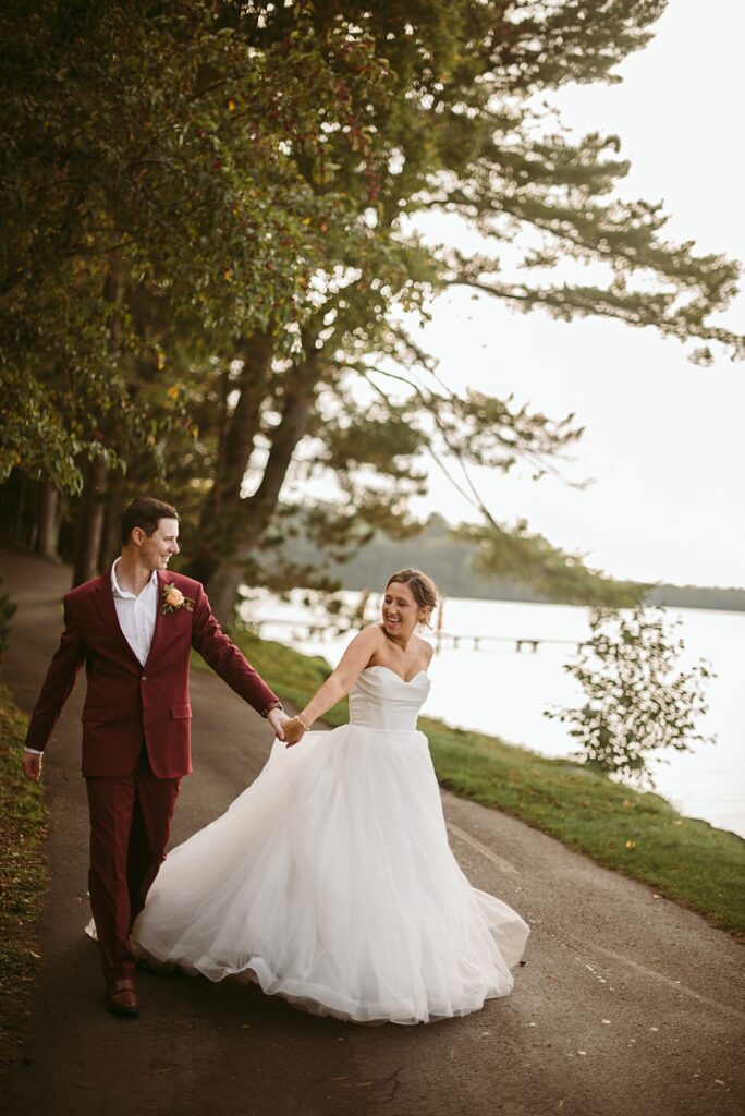 A bride in a white gown and a groom in a maroon suit walk hand in hand on a path by a lake, smiling at each other, surrounded by green trees—a perfect moment for their 2025 wedding recap.