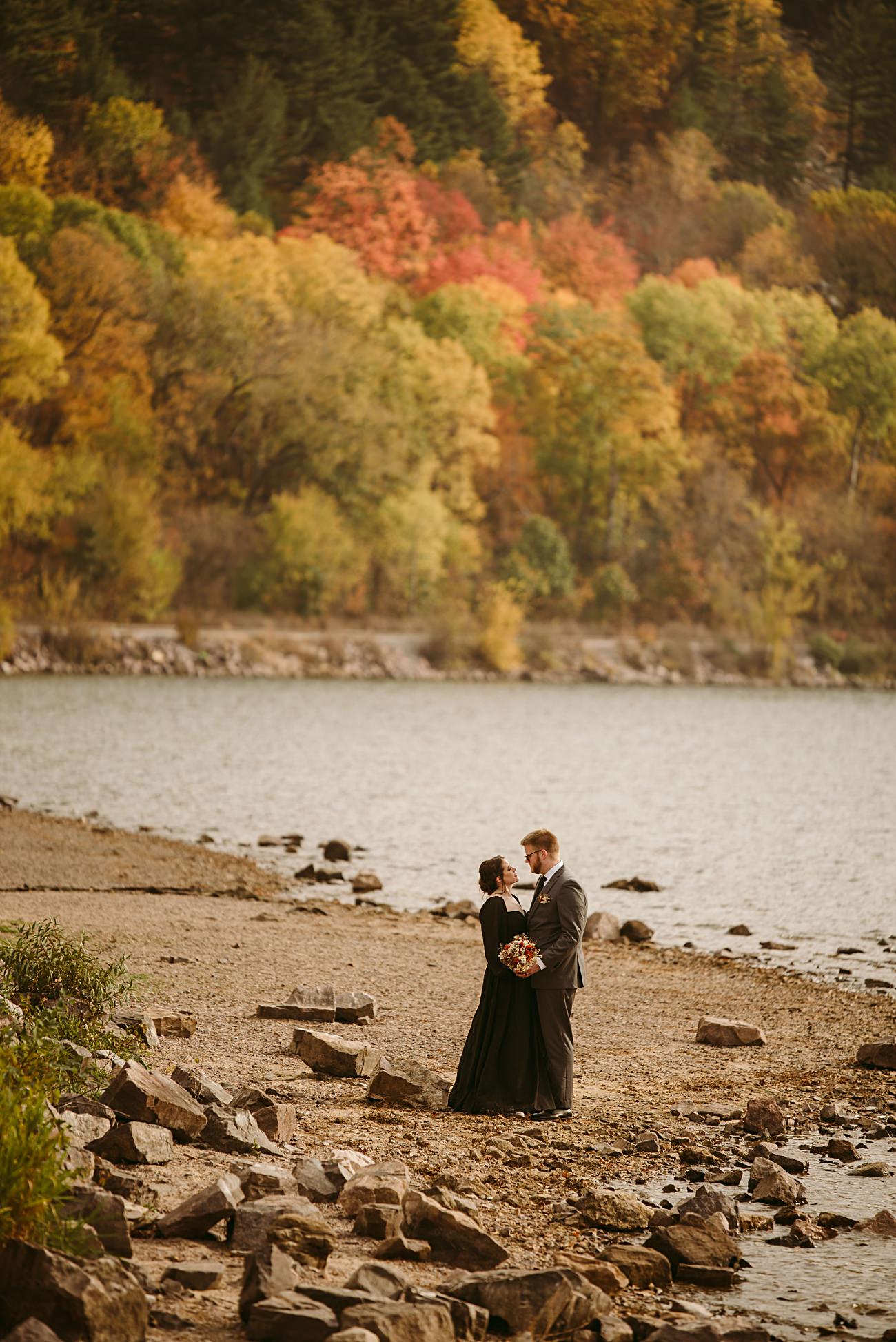 A couple dressed formally stands on a rocky lakeshore, gazing at each other with a bouquet in hand—capturing the magic of their 2025 wedding as vibrant autumn trees glow in yellow, orange, and red. Perfect for a heartfelt wedding recap.