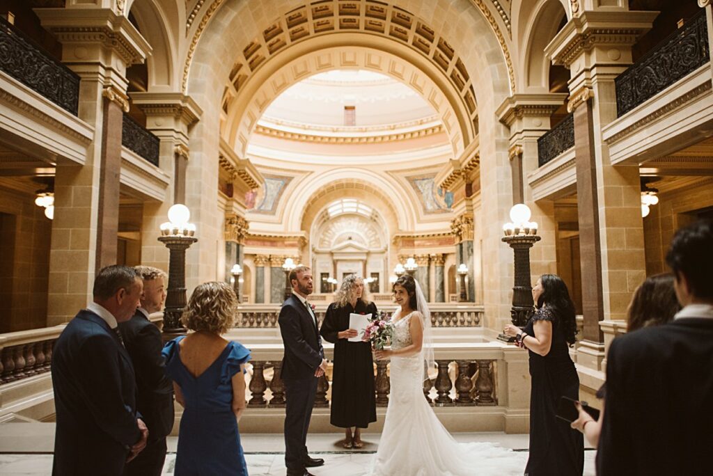 A couple stands facing each other in wedding attire during their 2025 Wedding inside a grand, ornate hall with marble floors, arched ceilings, and warm lighting, surrounded by a small group of guests.