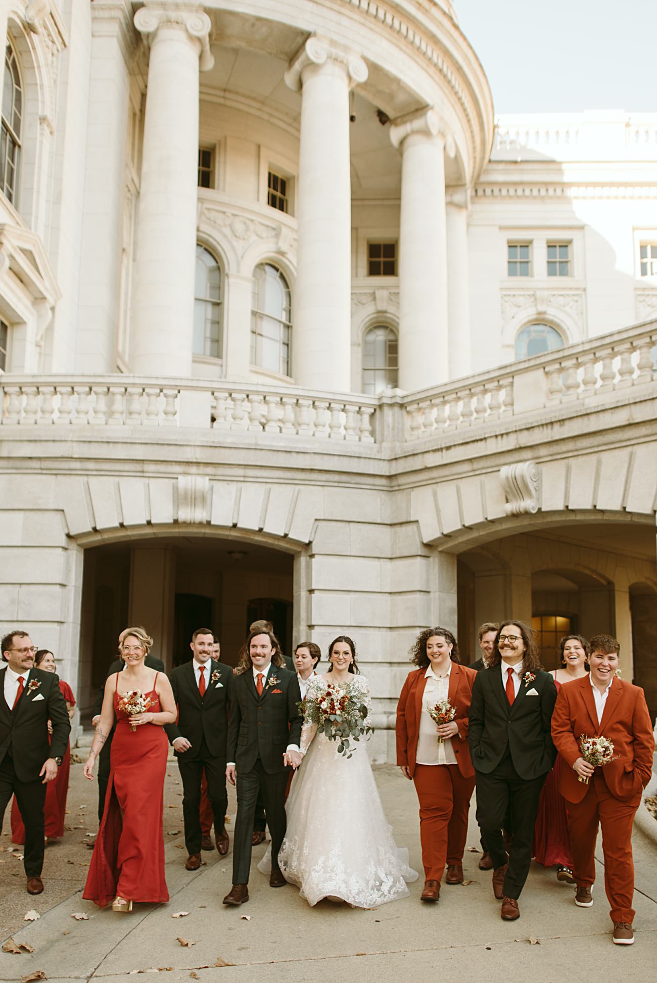 A bride and groom walk outside a grand, white stone building with their wedding party, dressed in red and black attire, smiling and holding bouquets. This joyful and elegant moment is perfect for a 2025 wedding recap under clear daylight.
