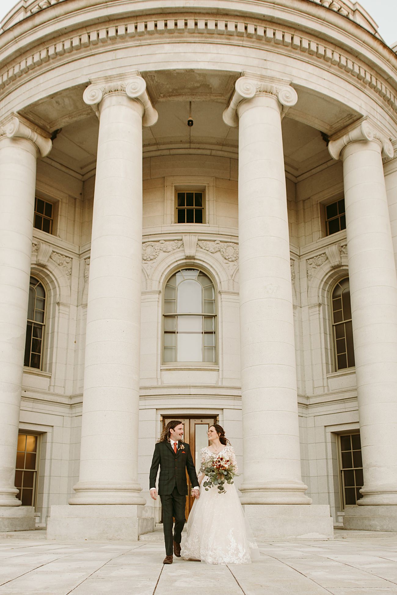 A bride in a white gown holding a bouquet and a groom in a dark suit walk hand in hand outside a grand building with tall white columns and arched windows, capturing a timeless moment perfect for any 2025 wedding recap.