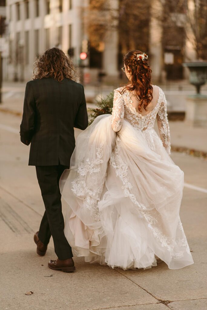A bride in a long, lace wedding gown with floral details walks beside a man in a dark suit on a city sidewalk, holding her dress to avoid the ground—a picture-perfect moment for any 2025 wedding recap, seen from behind as they stroll outdoors.