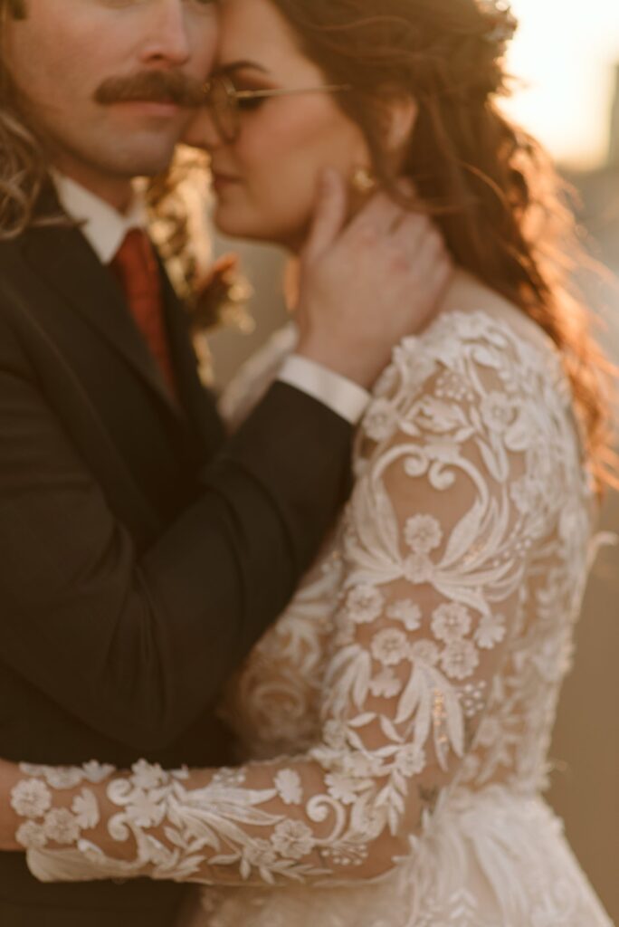 A couple in formal attire embraces closely, capturing a tender 2025 Wedding moment. The person in a white, floral lace dress has long hair, while the other, in a suit and tie with a mustache, stands together in softly focused, warm light.