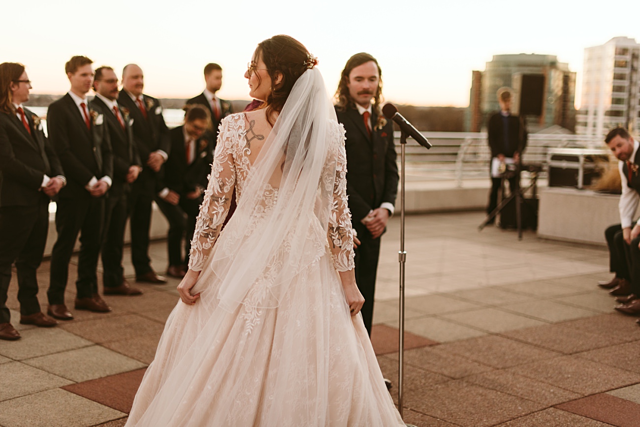 A bride in a lace gown with a veil stands facing her groom at an outdoor 2025 wedding ceremony, surrounded by groomsmen, with city buildings glowing in the sunset.