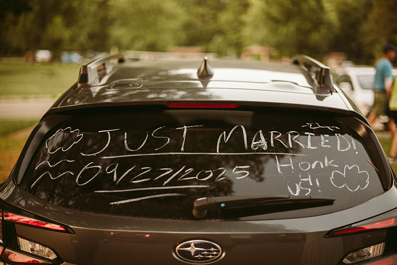 A car’s rear window is decorated with “JUST MARRIED!” and “09/27/2025” in white, with clouds, waves, and “Honk 4 us!”—the perfect snapshot for a 2025 Wedding recap in a scenic park setting.