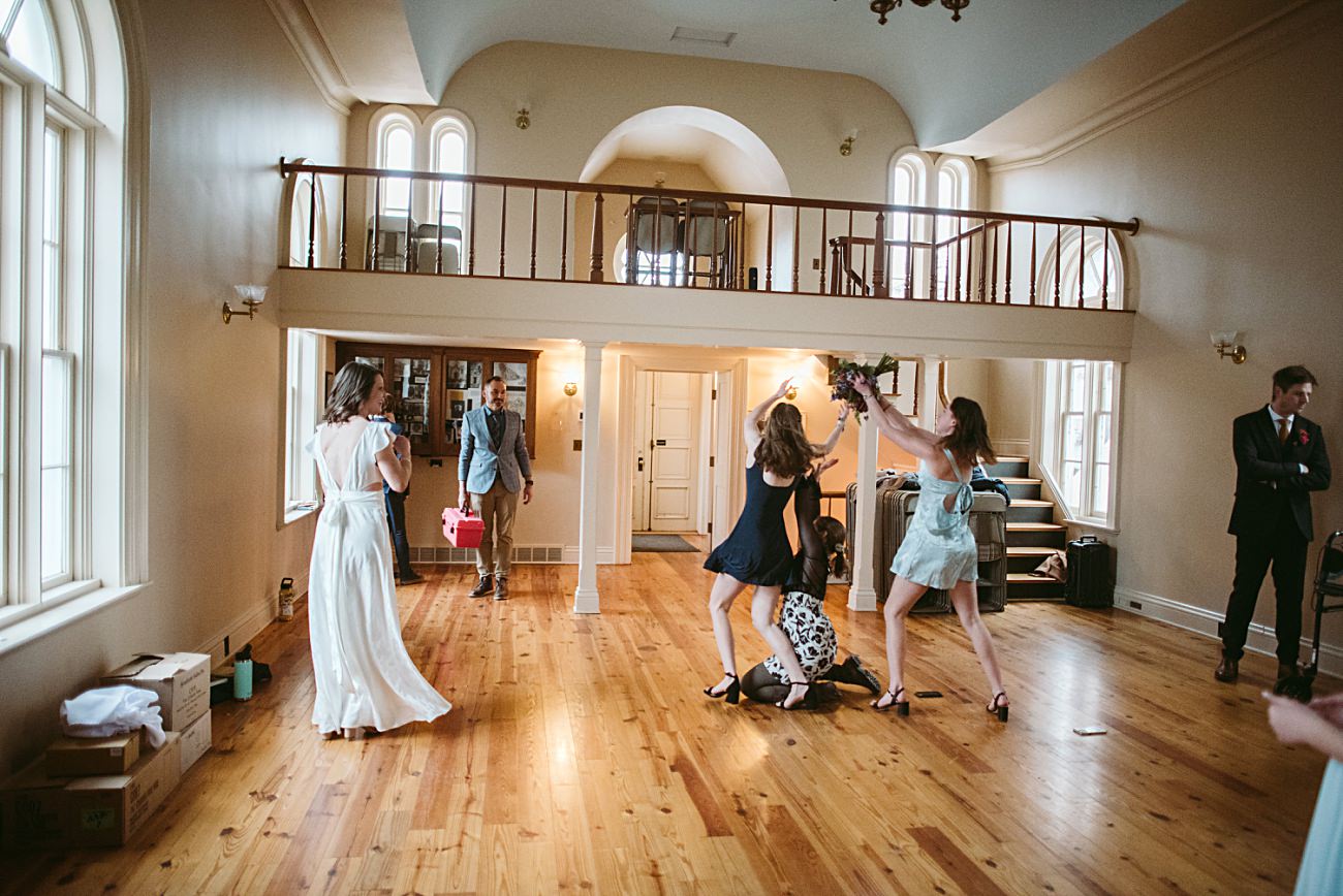 Several women in dresses playfully compete for a bouquet in the center of a bright, spacious room with wooden floors during this joyful 2025 wedding. Other guests watch the scene, while boxes are scattered on the floor—perfect for any wedding recap.