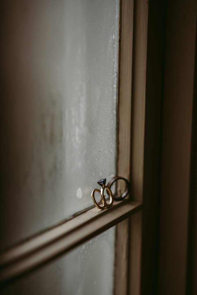 A close-up of three rings, including a diamond engagement ring, resting on the wooden sill of a frosted window. The softly blurred background adds a moody touch—perfect for capturing 2025 wedding highlights in an intimate atmosphere.