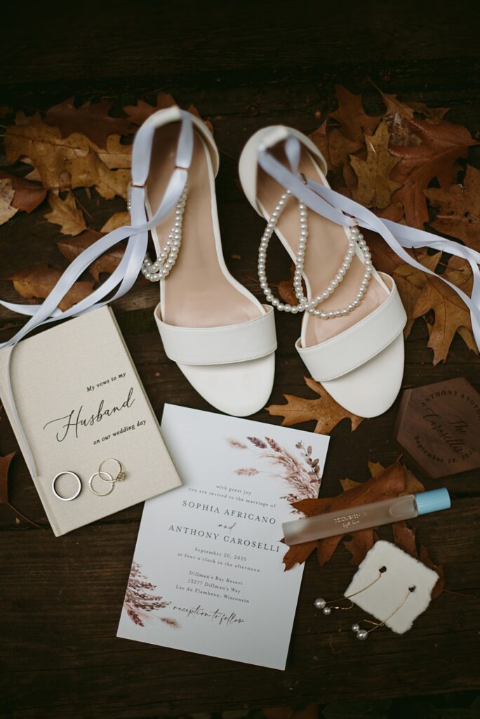 Flat lay of 2025 wedding items on a wooden surface with autumn leaves: white heels with pearls, a wedding invitation, rings in a box, perfume, earrings, and a personalized leather coaster—perfect for your wedding recap.