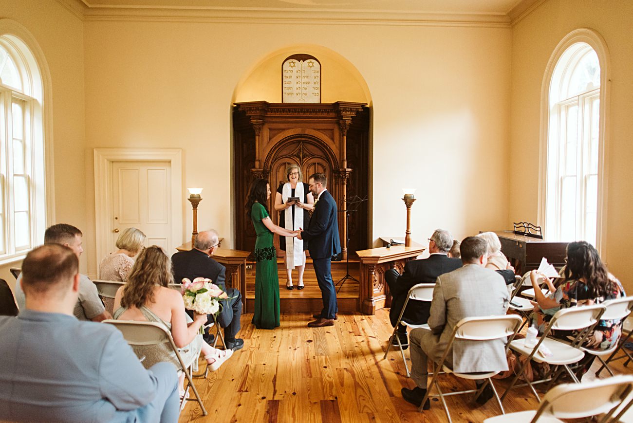 A couple stands holding hands at the altar in a small chapel, facing an officiant. Guests watch the 2025 wedding ceremony from white chairs in a warmly lit room with wooden floors and large windows.