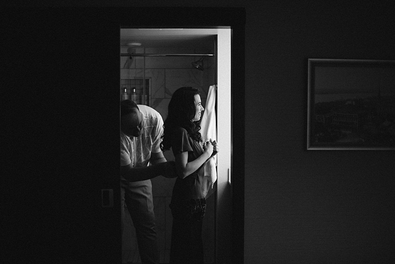 A man helps a woman zip up her dress in a brightly lit bathroom while the rest of the room stays dark. The woman smiles, facing the light—a timeless black-and-white moment perfect for a 2025 wedding recap.
