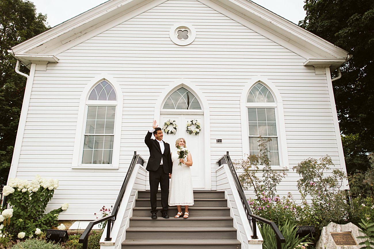 A bride and groom stand on the steps of a white wooden chapel. The groom waves while the bride, radiant with her bouquet, smiles. Flowers and greenery surround them in this picturesque 2025 wedding moment.