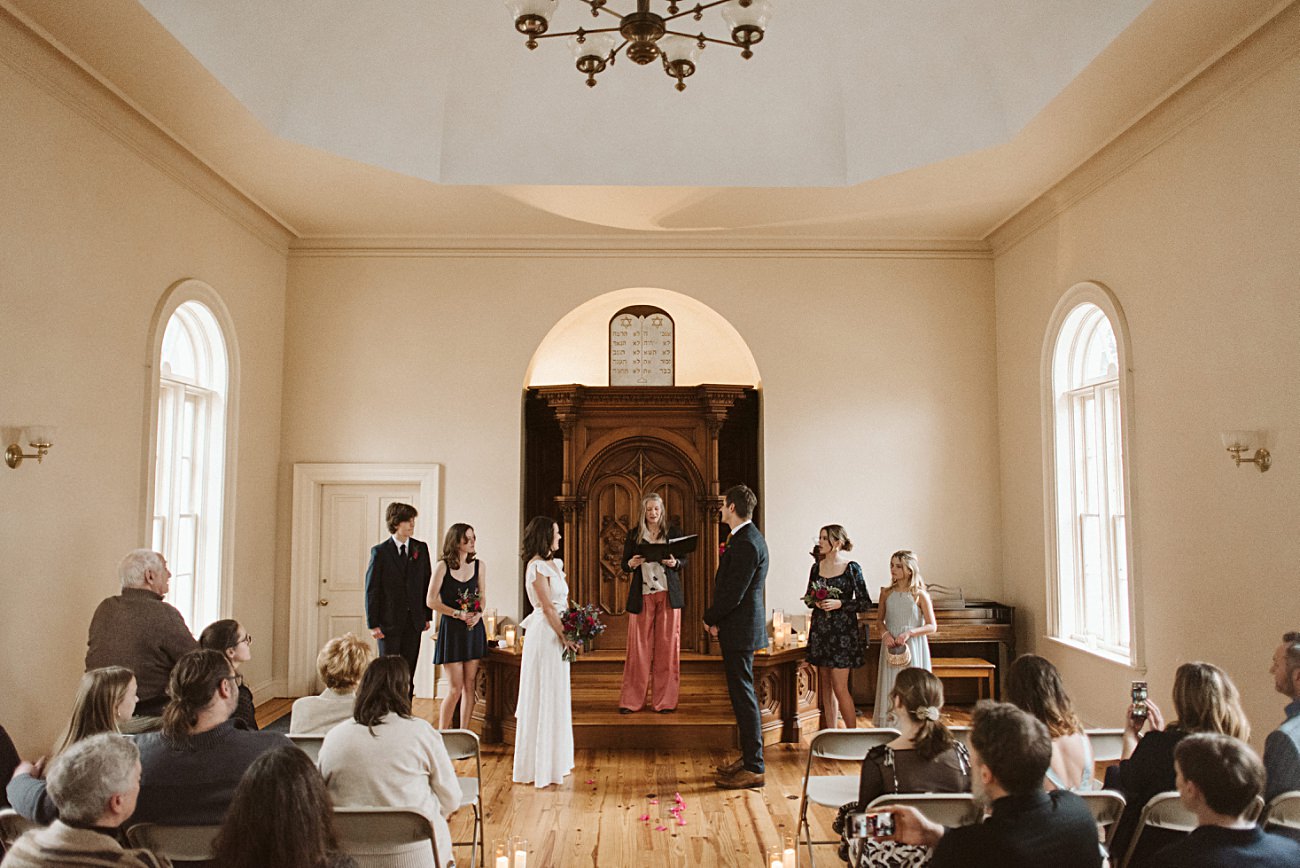 A couple stands at the altar during a wedding ceremony in the light-filled Gates of Heaven chapel in Madison, Wisconsin. Guests are seated, attendants stand on either side, and the intimate atmosphere is filled with joy.