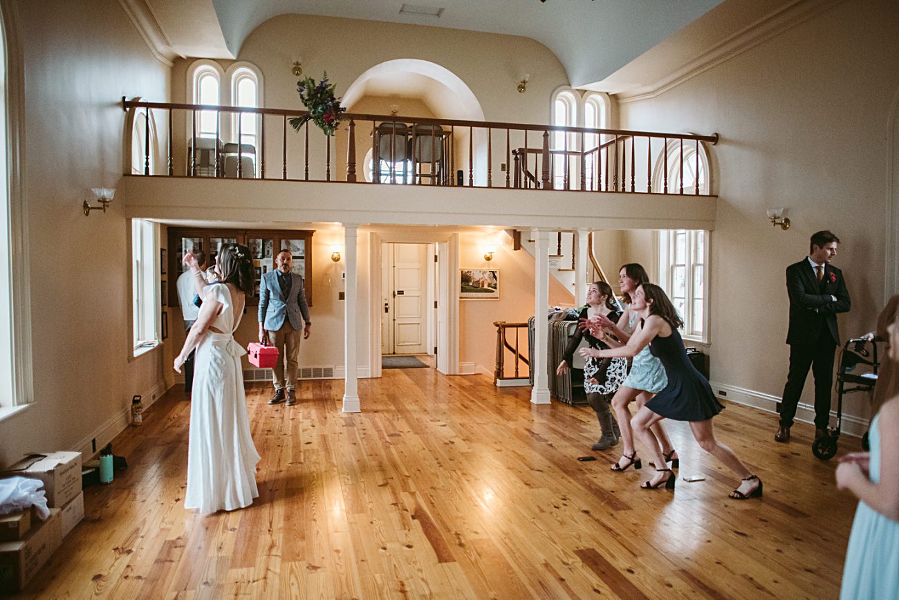 A bride in a white dress stands on a wooden floor at Gates of Heaven in Madison, Wisconsin, preparing to toss her bouquet as several women eagerly reach for it. A few people watch from the side in the spacious, light-filled room with a loft.