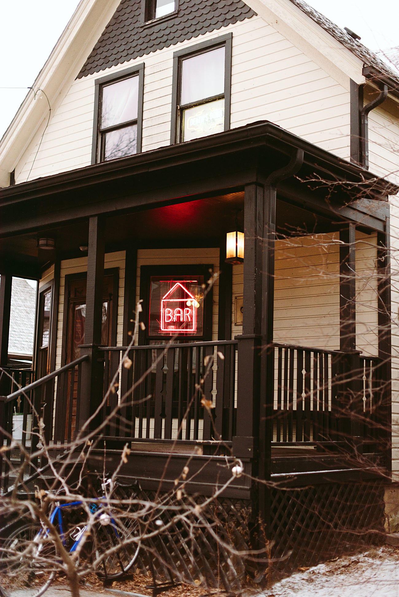 A two-story house with white siding and dark trim has a front porch displaying a red neon BAR sign shaped like a house. In the yard, bare branches frame a blue bicycle, evoking Madison Wisconsin charm on a quiet evening.