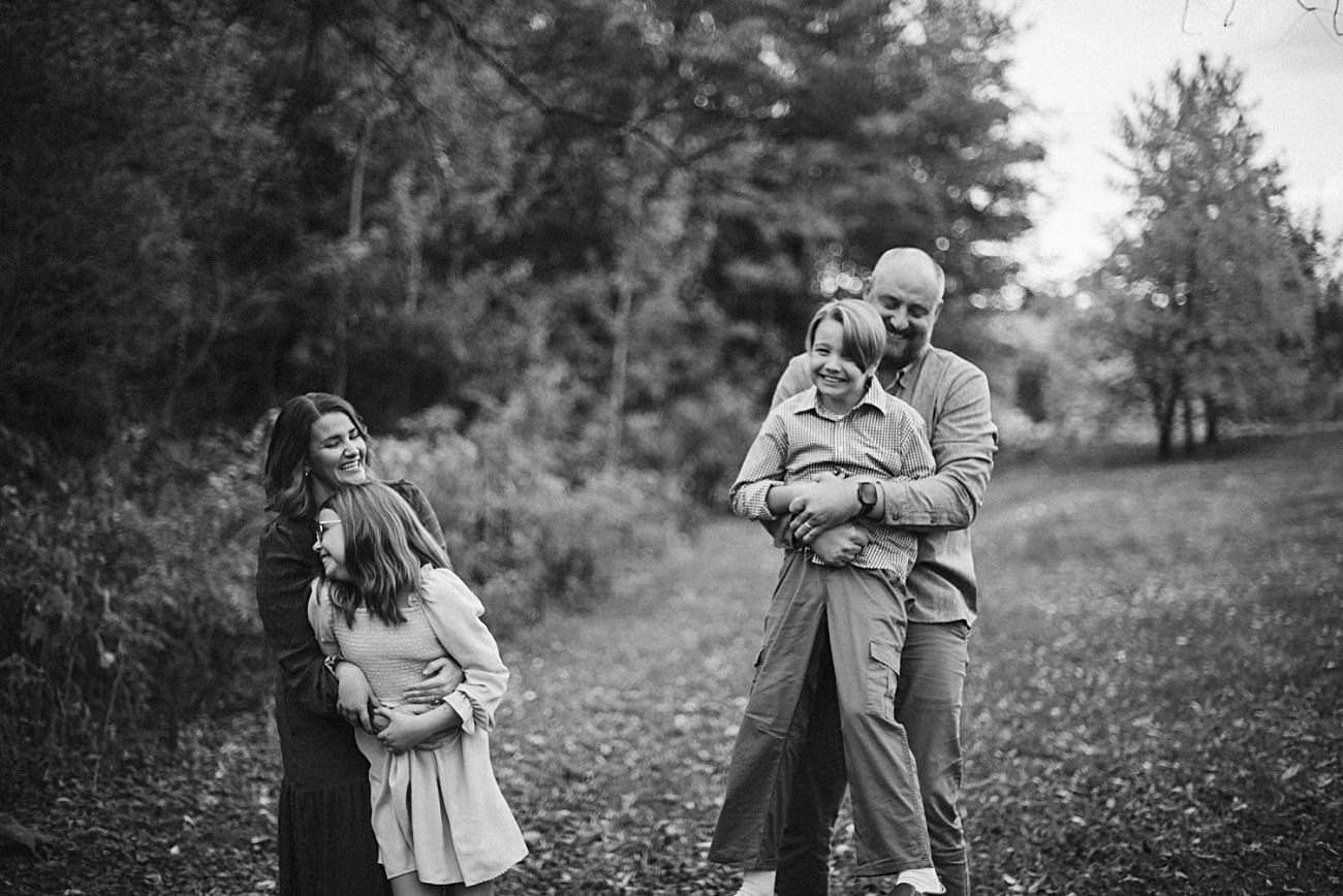 A black-and-white photo of a smiling family outdoors. Perfect for Wisconsin family photos, it captures a woman hugging a young girl on the left and a man with another laughing girl on the right, standing on a leafy path with trees behind them.