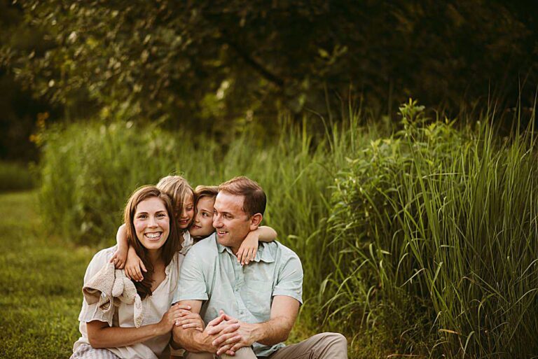 A smiling family of four sits close together in tall grass outdoors for their 2025 Family Photos. The parents sit in front, while their two children hug them from behind. The green setting features Wisconsin trees in the background.