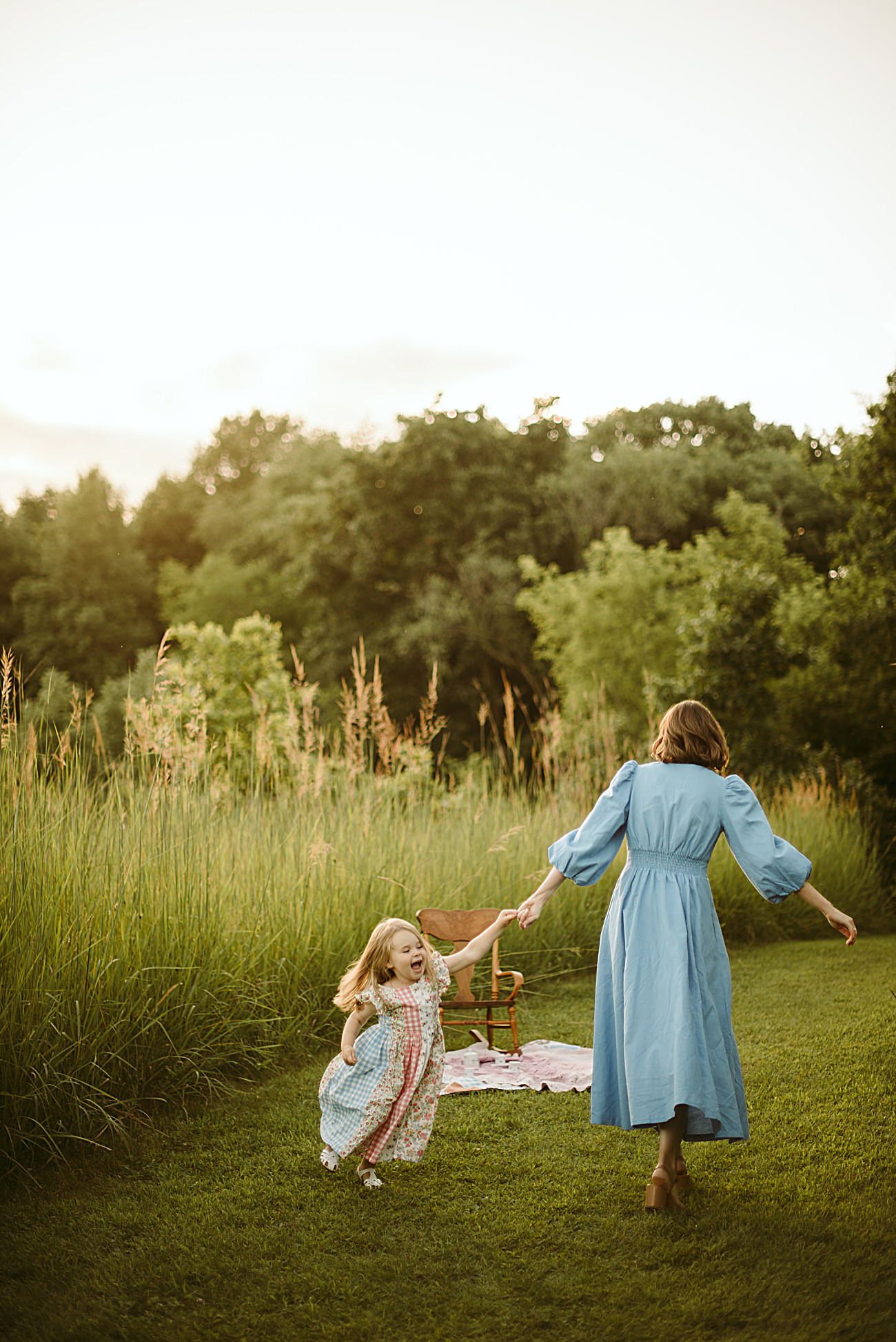 A woman in a blue dress holds hands with a young girl in a patterned dress as they walk through a grassy field at sunset—capturing the warmth of Wisconsin Family Photos for their 2025 Year in Review.