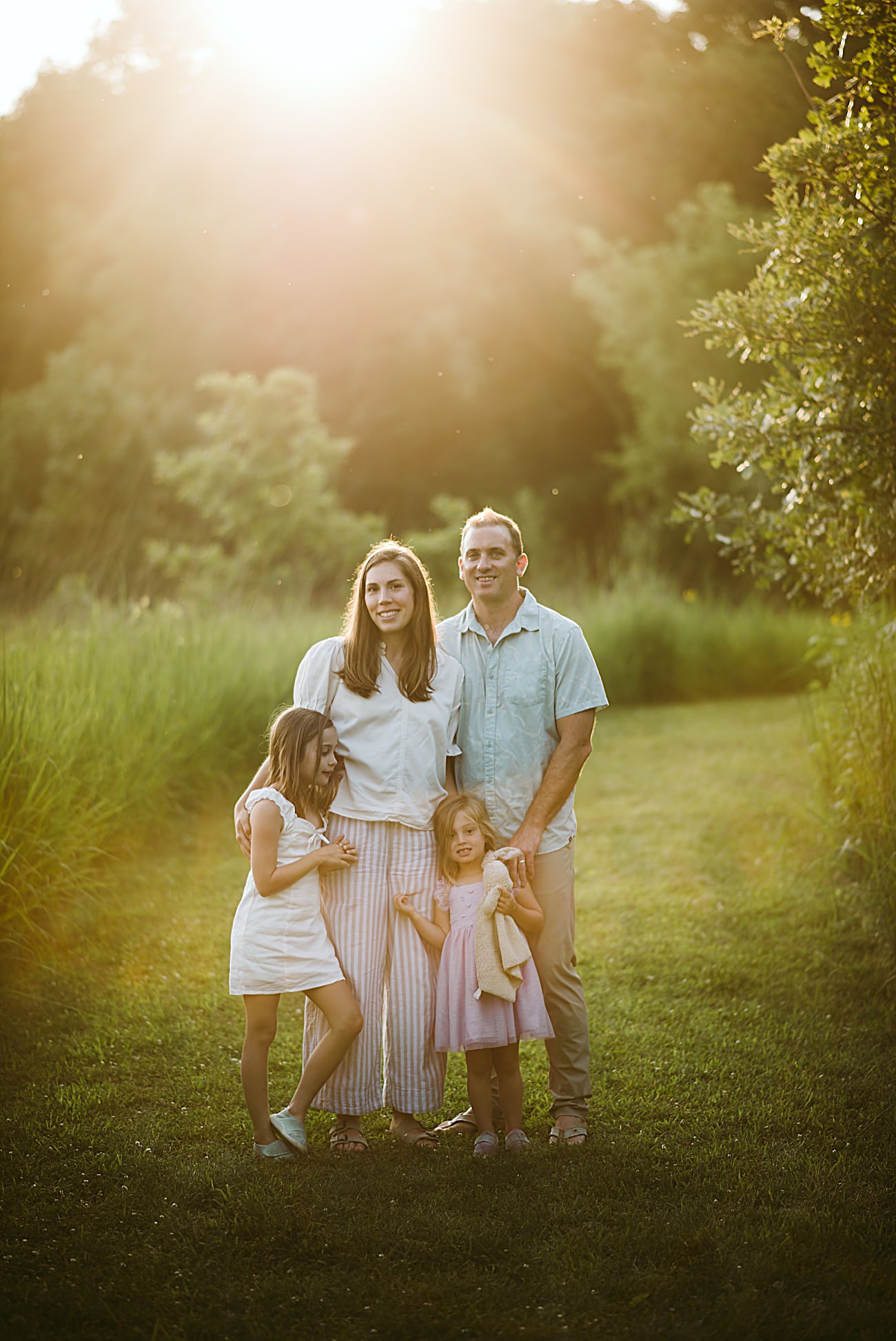A Wisconsin family of four—two adults and two young girls—stand together on a grassy path in a sunlit field, surrounded by greenery, with warm sunlight shining behind them for their 2025 Wisconsin family photos.