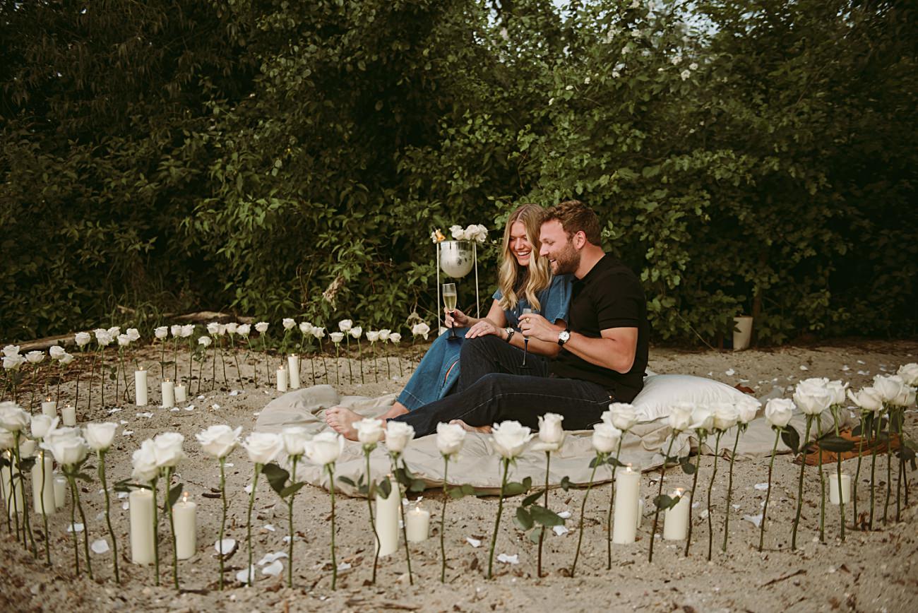 A couple sits on a round blanket surrounded by white roses and candles on sand, smiling and holding drinks, celebrating a magical proposal with lush greenery in the background.