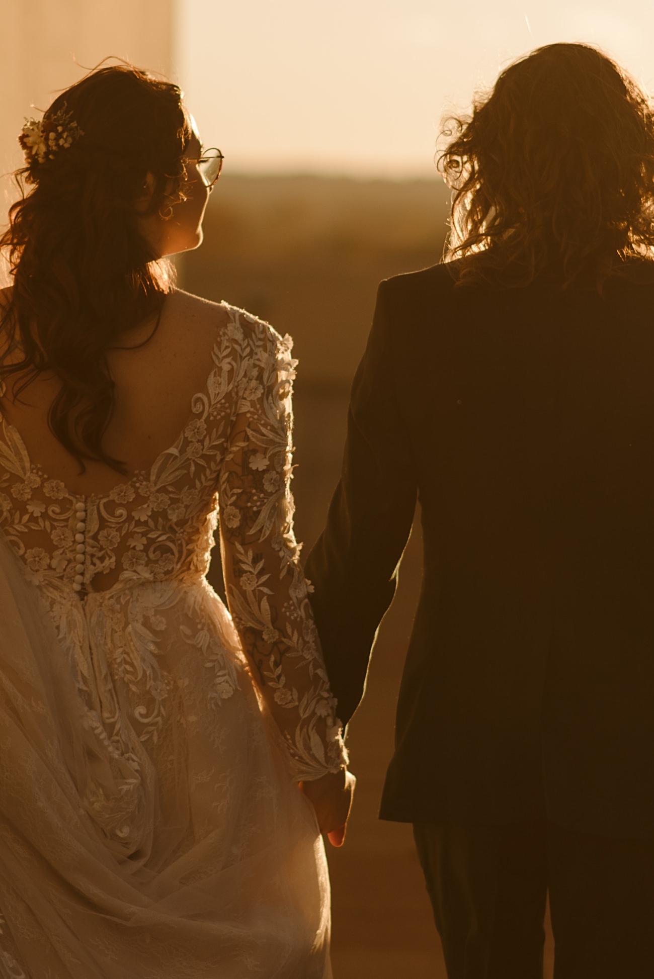A bride in a lace wedding dress and a groom in a dark suit stand hand in hand, viewed from behind, bathed in warm, golden sunlight at their Blue Hour Rooftop Wedding atop Monona Terrace in Madison, Wisconsin.