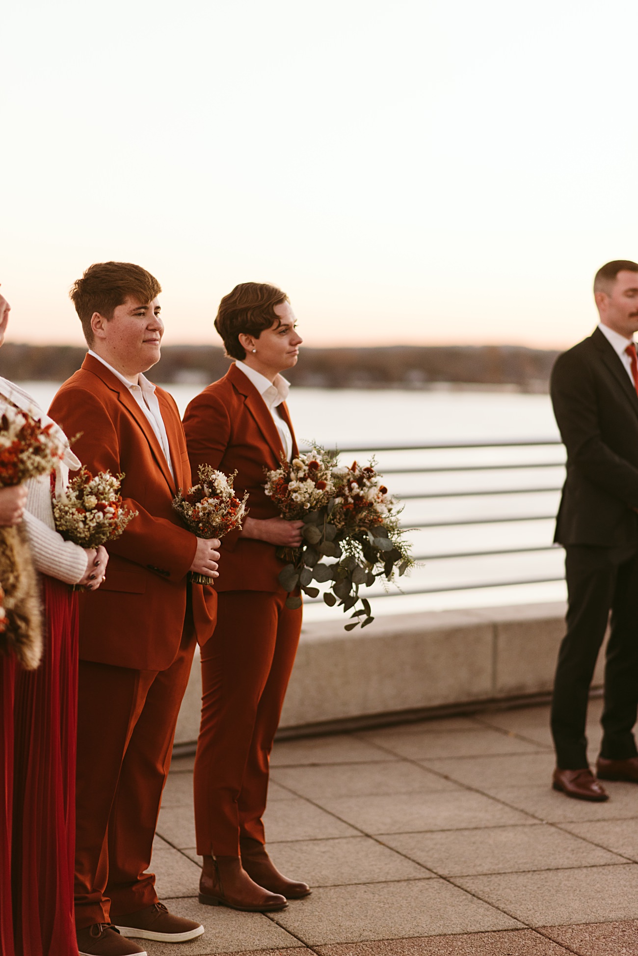 Two people in matching rust-colored suits hold bouquets while standing outdoors at sunset on Monona Terrace in Madison, Wisconsin. A body of water and distant hills create a stunning backdrop, with others partially visible beside them.