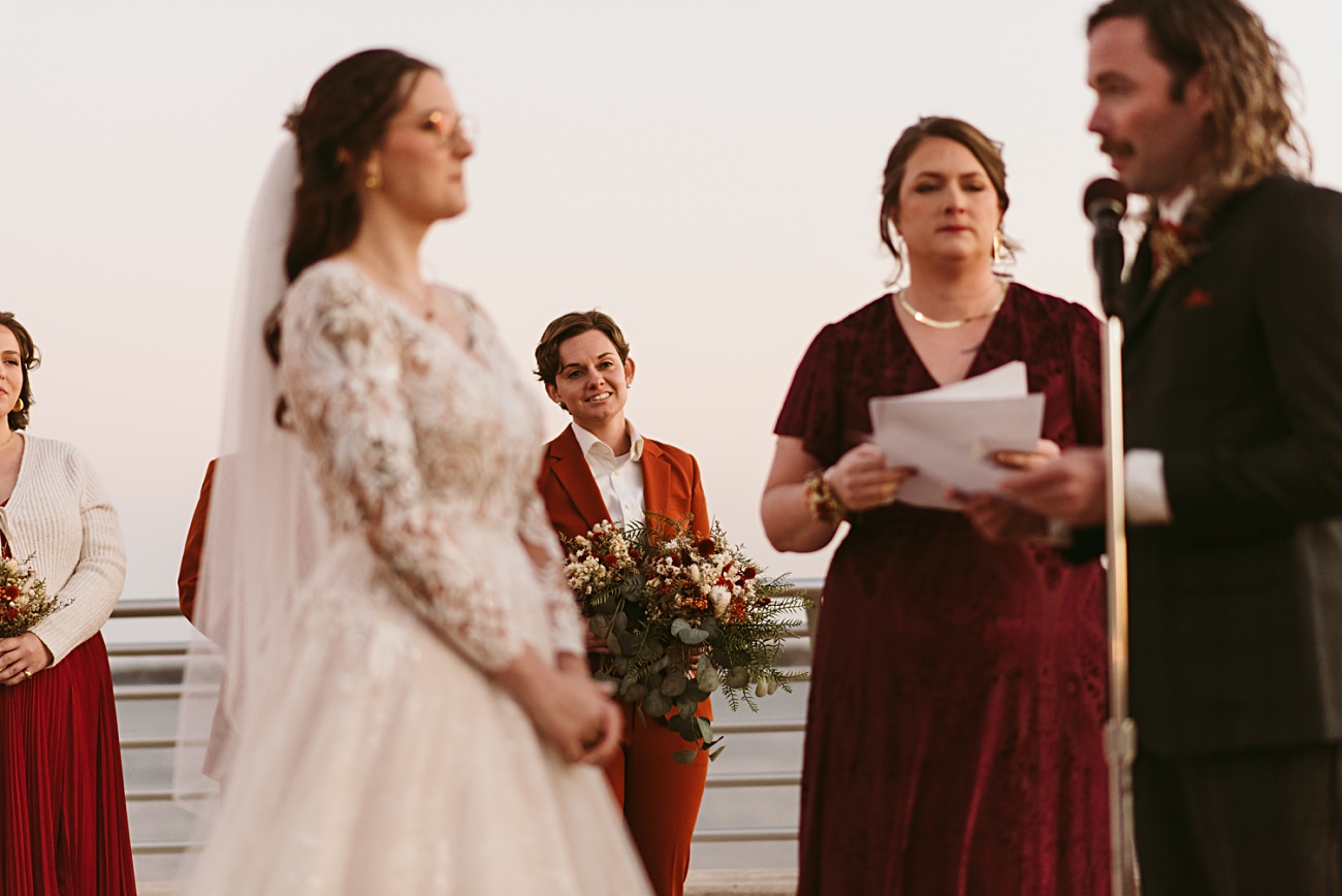 A Blue Hour Rooftop Wedding at Monona Terrace in Madison, Wisconsin: a bride in a white gown stands with attendants as someone reads at a microphone; a smiling person in a red suit holds a bouquet in the background under the sunset sky.
