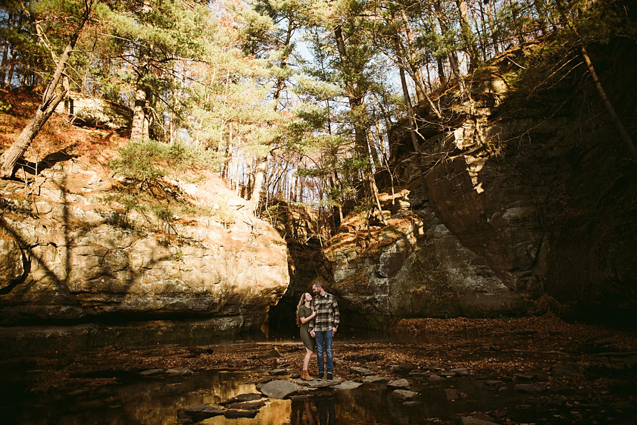 A couple stands together on rocks near a shallow stream, surrounded by tall cliffs and sunlit trees in a forested Wisconsin canyon—one of the best places for engagement photos.