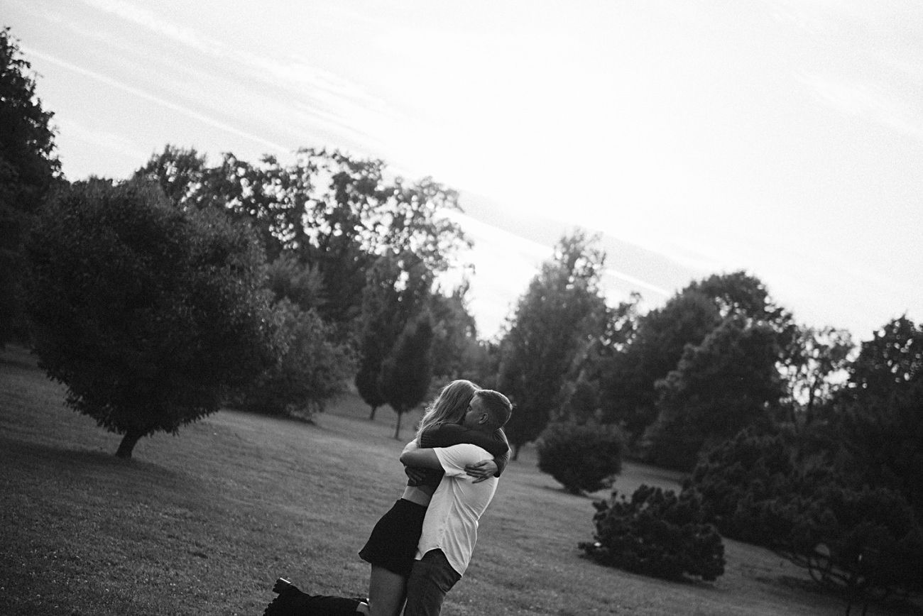 A couple embraces and kisses in a park surrounded by trees, with sunlight filtering through the background. Captured in black and white at the UW Arboretum, Madison, the image exudes a romantic and serene atmosphere.