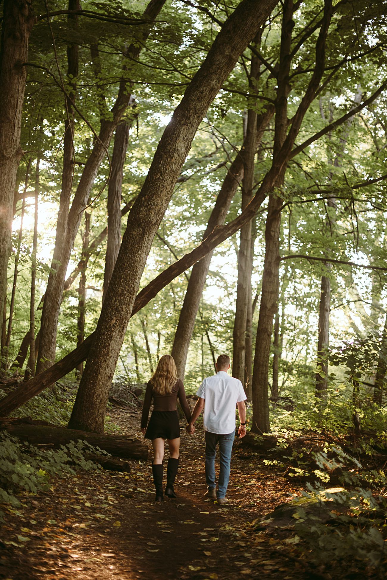 A couple holding hands walks along a dirt path through the lush, green UW Arboretum in Madison, sunlight streaming through the trees—an idyllic spot for a proposal.