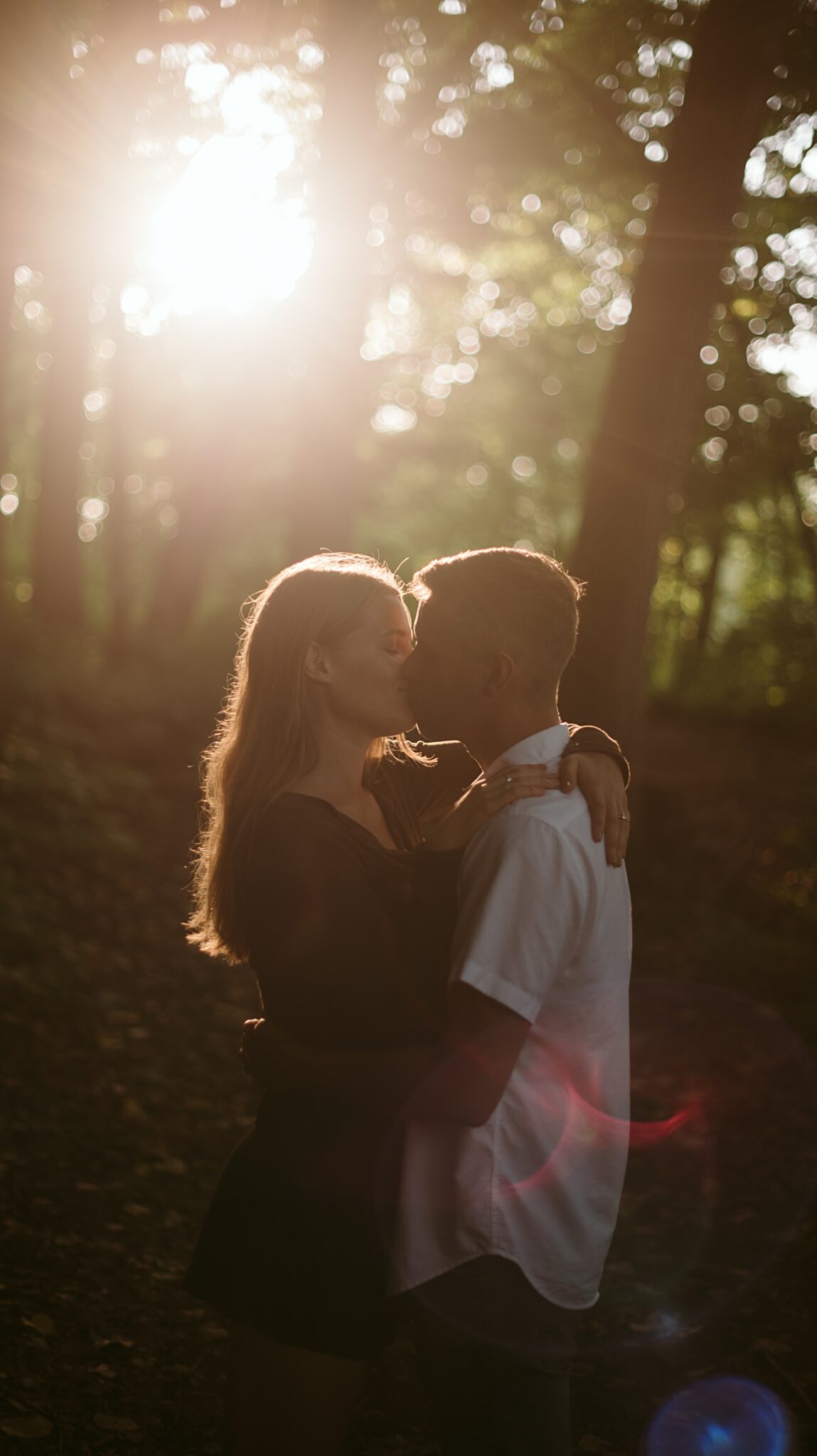A couple embraces and gently kisses in a sunlit forest at the UW Arboretum in Madison, with soft sunlight streaming through the trees, creating a romantic and dreamy proposal atmosphere.