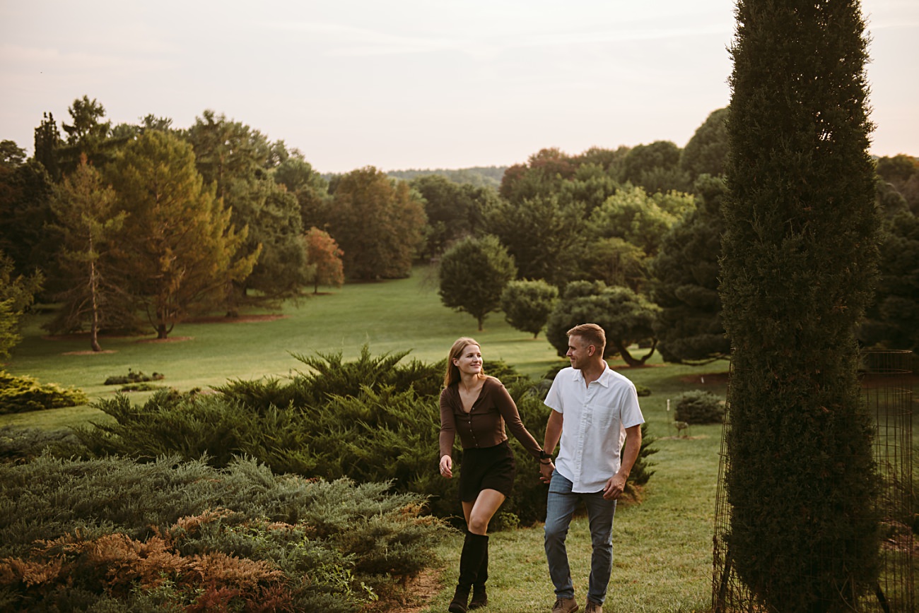 A couple walks hand in hand through the lush green UW Arboretum in Madison, smiling at each other on a warm, sunny day as they celebrate their proposal.