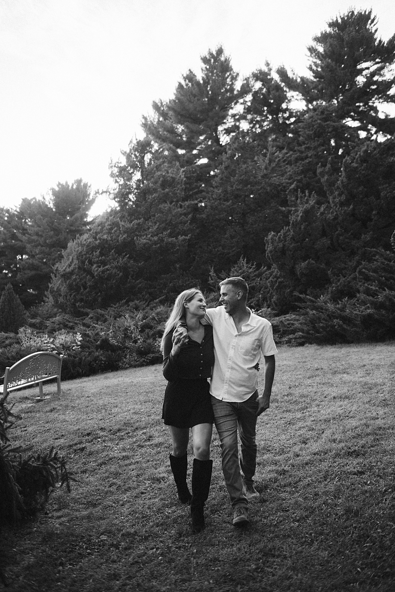 A couple walks closely together on a grassy lawn, smiling at each other amid tall trees and shrubbery in a natural outdoor setting. This black and white image captures the charm of a UW Arboretum proposal in Madison.