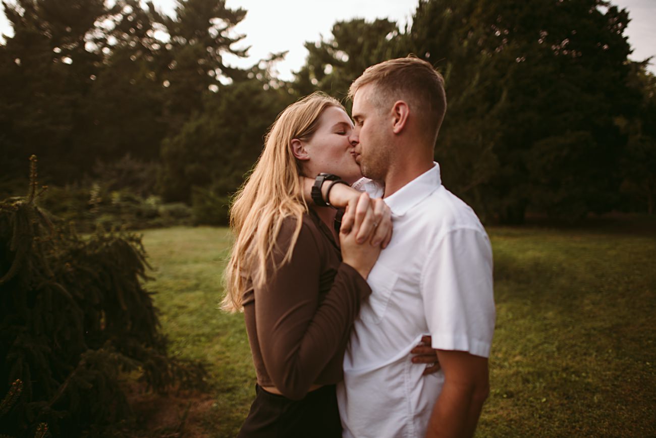 A couple stands outdoors in a grassy area with trees at UW Arboretum in Madison, sharing a kiss while holding each other closely. The woman has long blonde hair and a brown top; the man has short hair and wears a white shirt. It’s a sweet proposal moment.