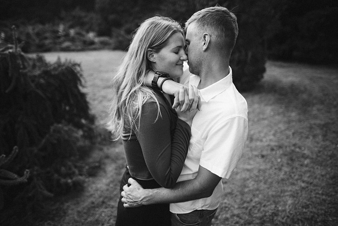 A black and white photo of a couple embracing outdoors at the UW Arboretum in Madison. The woman smiles with her eyes closed while the man holds her close in a grassy area, trees blurred in the background—a perfect proposal moment.