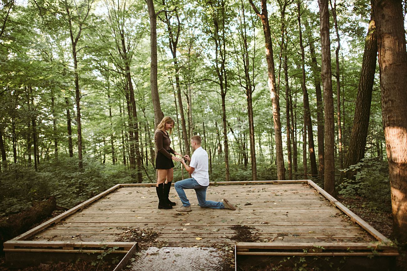 A man kneels on a wooden platform in the UW Arboretum in Madison, holding a ring box and proposing to a woman, who stands facing him and looks surprised. Tall trees surround the platform, bathed in natural daylight.
