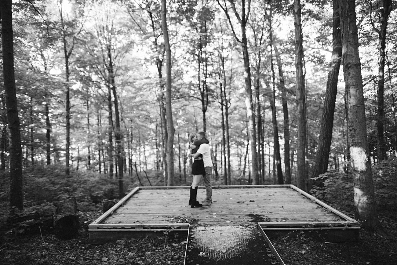 A couple stands on a small wooden platform in the UW Arboretum, Madison, embracing each other. The black and white scene is surrounded by tall trees, with dappled sunlight filtering through the leaves—a perfect proposal moment.