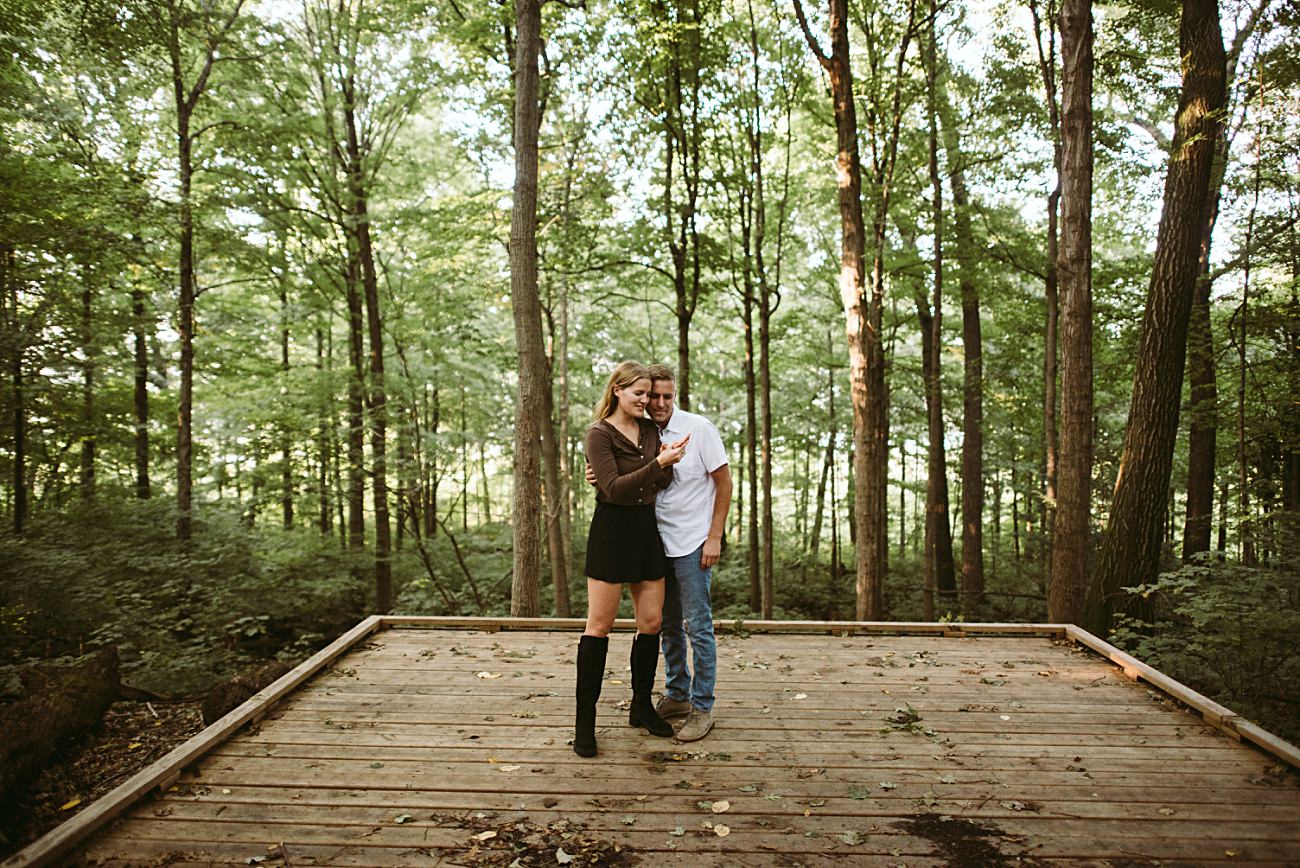 A woman in a black dress and boots and a man in a white shirt and jeans stand smiling together on a wooden platform at the UW Arboretum in Madison, surrounded by tall green trees—the perfect spot for a proposal. The woman is pointing and laughing.