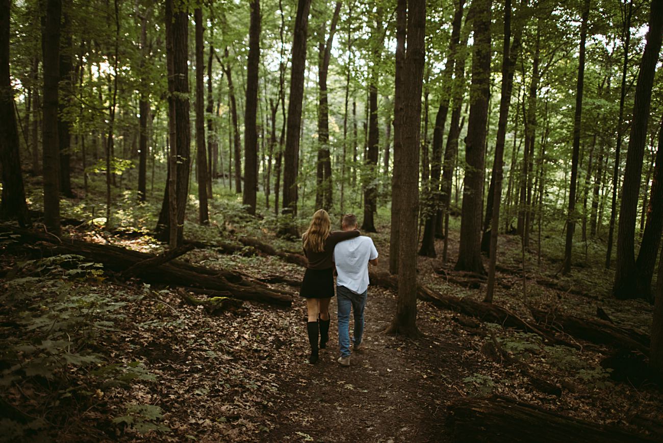 A couple walks arm in arm along a dirt path through the lush, green UW Arboretum in Madison, surrounded by tall trees and scattered fallen logs as sunlight filters through the leaves above.