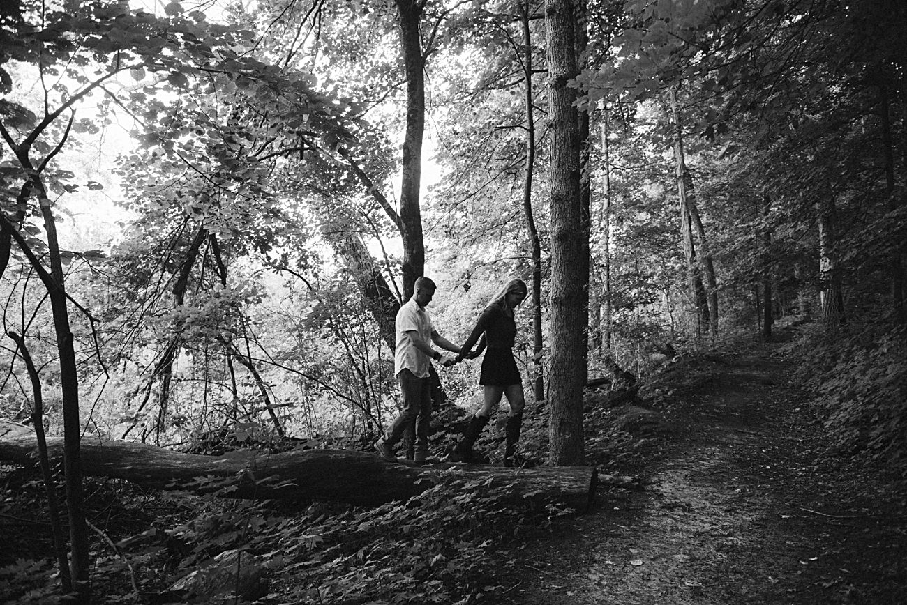 A black and white photo of two people walking along a forest path in the UW Arboretum, Madison, holding hands as they carefully step over a fallen tree surrounded by dense foliage and tall trees—a perfect proposal spot.