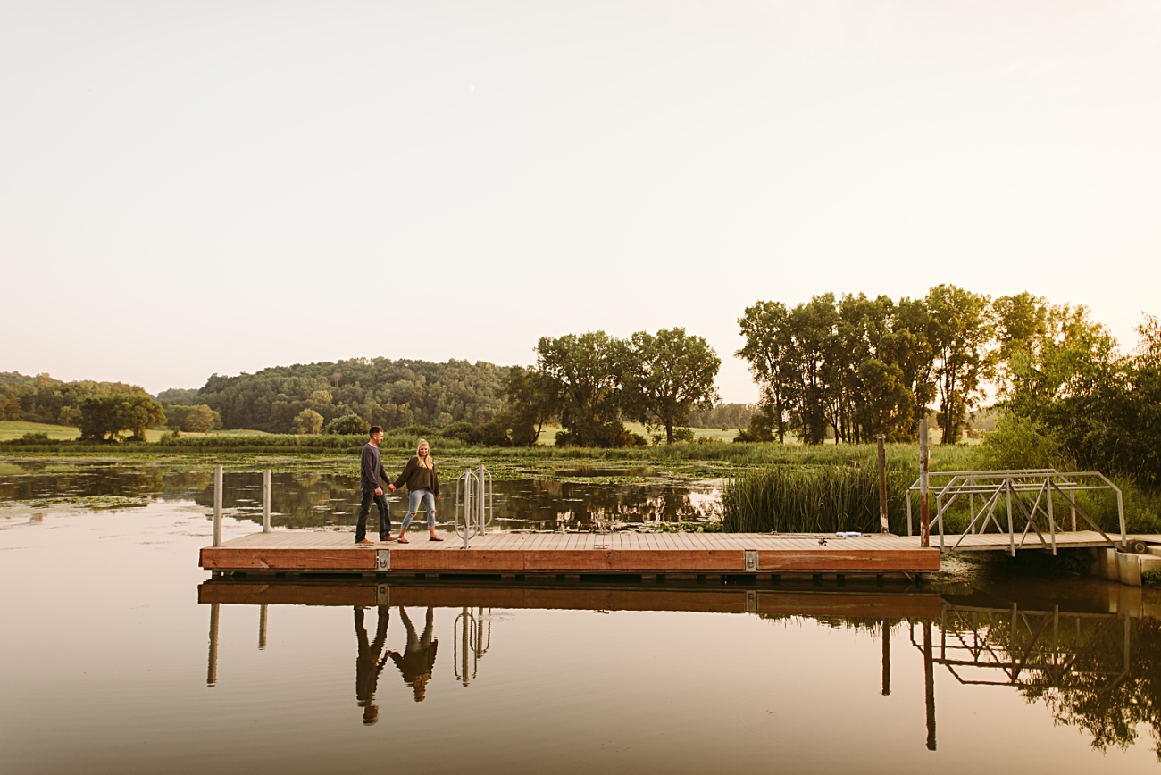 A couple sits together on a wooden dock at Middleton Tree Farm over a calm lake, surrounded by green trees and grassy hills at sunset—the perfect setting for an Engagement Session, with warm light reflecting on the water.